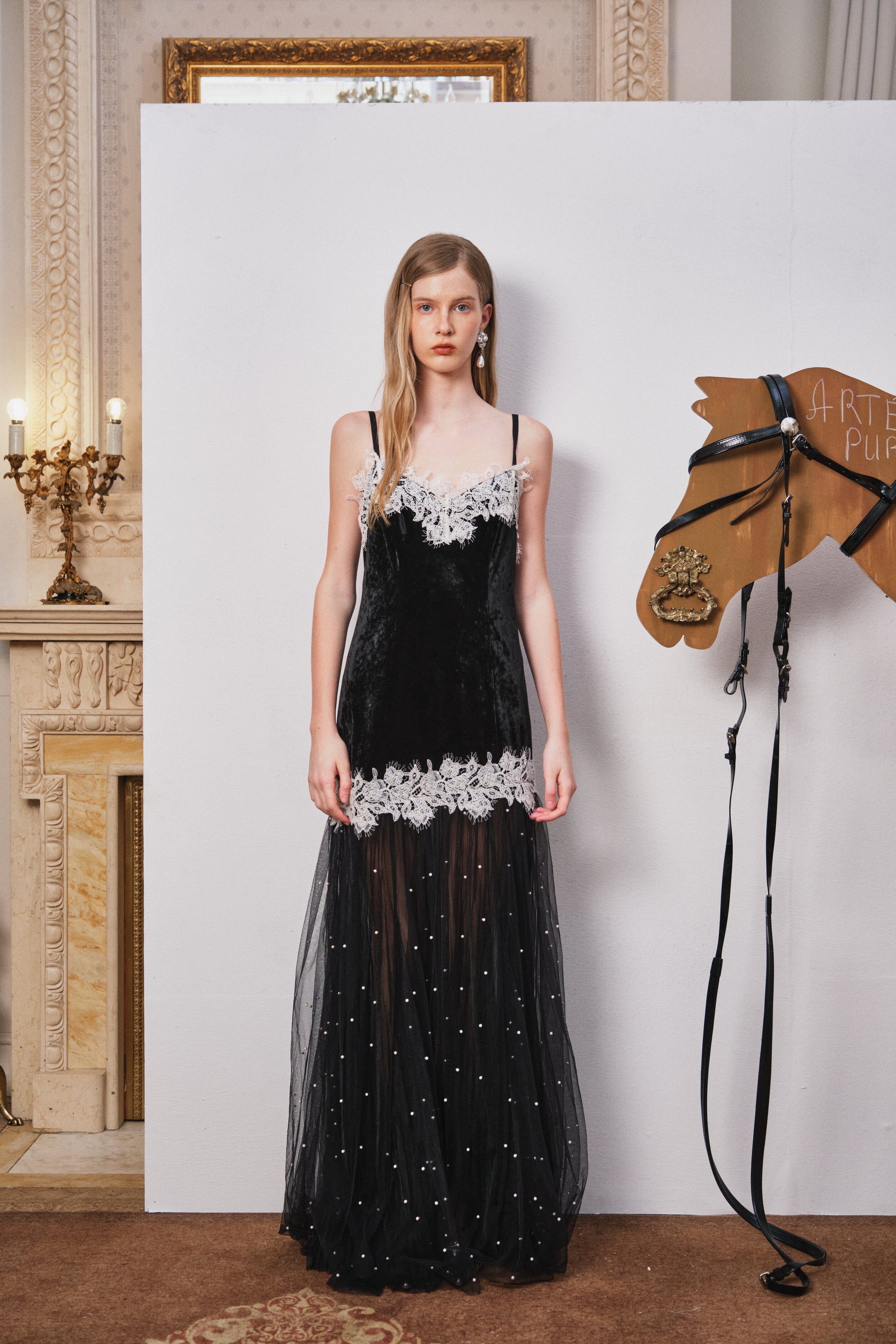 Woman in a black evening gown with white lace details standing next to a horse head sculpture in an elegant interior setting.