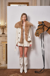 Woman in a fur-trimmed coat standing next to a wooden horse figure in an elegant room.