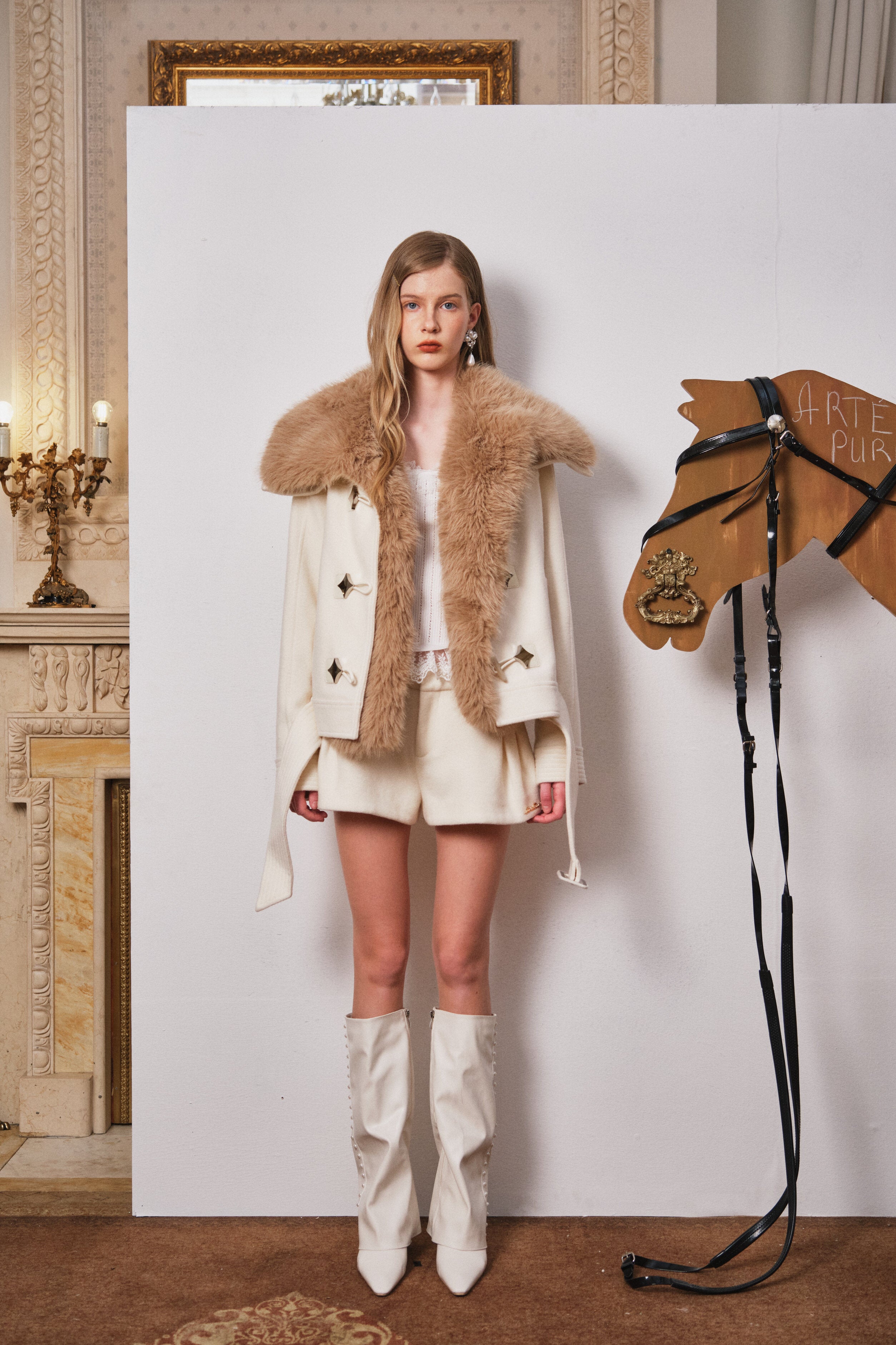 Woman in a fur-trimmed coat standing next to a wooden horse figure in an elegant room.