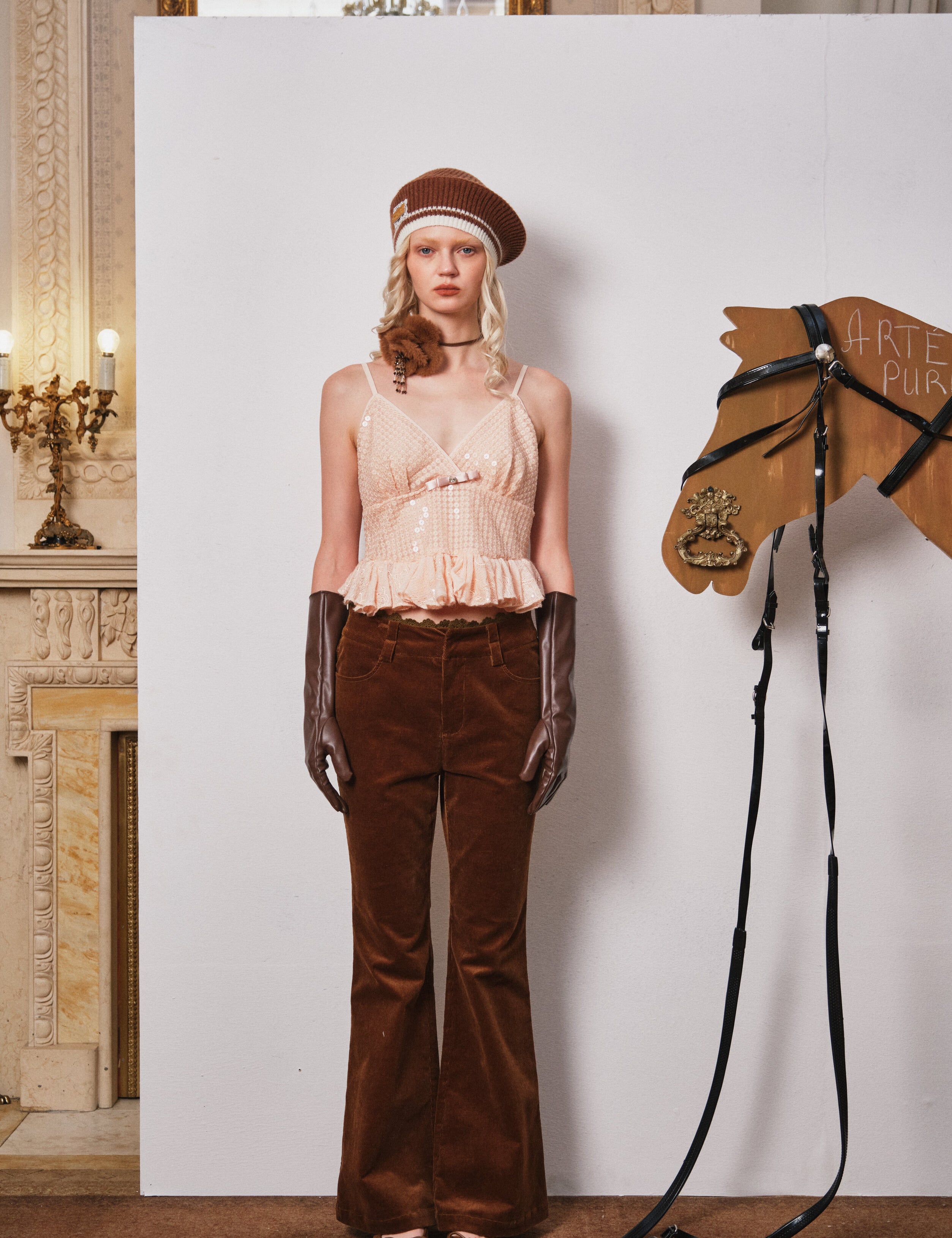 Woman in vintage outfit with horse head sculpture and bridle against a white wall.